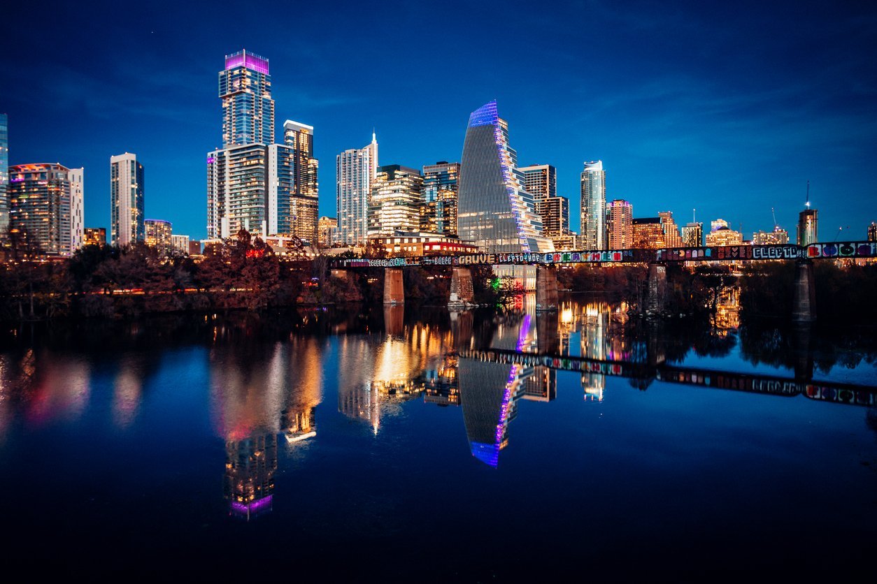 Nighttime city skyline across a calm river, neon-lit buildings reflected in the water, with a graffiti-covered bridge in the foreground