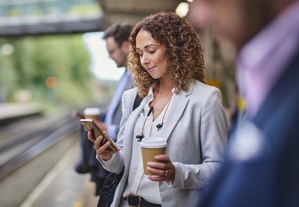 Businesswoman at a train platform checks her smartphone while holding a coffee cup to go.