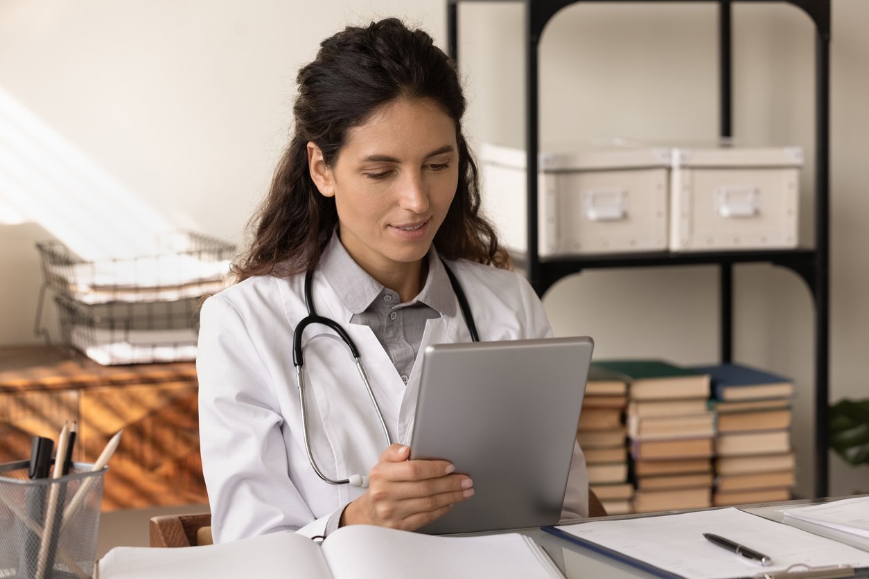 Female clinician in a white coat with a stethoscope reads a tablet at a desk in a medical office.